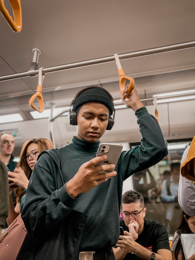 A person on a train, holding onto the ceiling handle while looking into their phone, and the people in the background on the train, all looking into their phones.