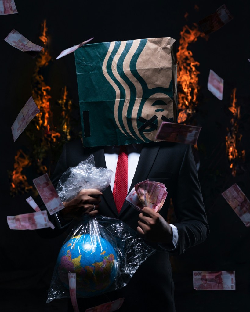 An image of a person in a black suit, white shirt, and red tie, holding cash in their hands with a Starbucks bag on their head. In the background is some fire and cash falling down.
