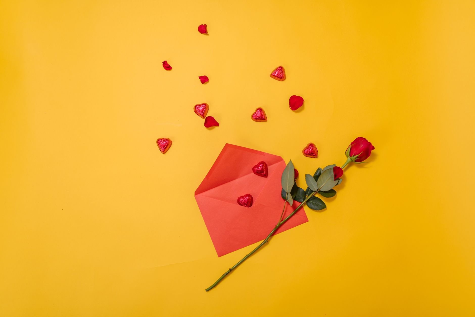 an open red envelope on a yellow background, with red love heart petals scattered coming out of the envelope and a single red rose places on top